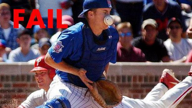 Ein Baseballspieler fängt einen Ball mit seinem Gesicht, während ein anderer Spieler versucht, die Homebase zu erreichen.