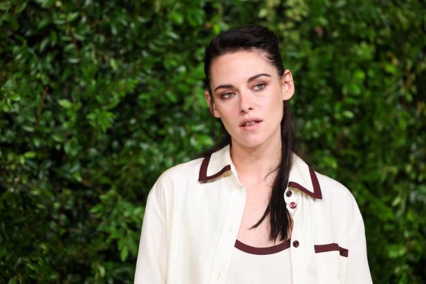 Kristen Stewart trägt ein cremefarbenes Hemd mit braunen Akzenten vor einer grünen Hecke.