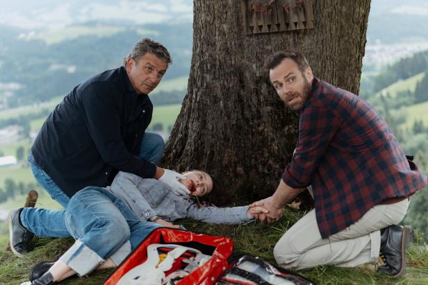 Zwei Männer knien neben einer verletzten Frau mit blutigem Mund an einem Baum und leisten Erste Hilfe.