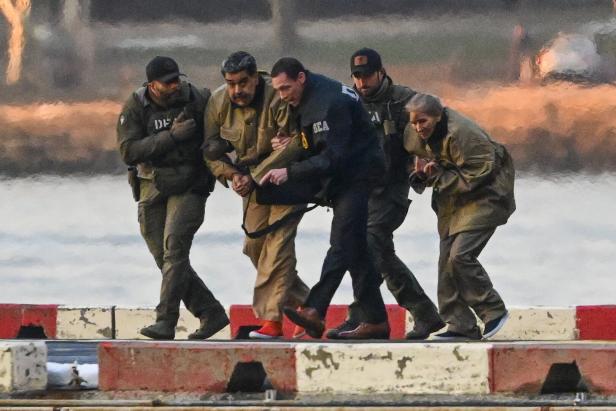 Fünf Personen in Schutzkleidung und Uniformen unterstützen sich gegenseitig beim Gehen auf einer nassen Straße.
