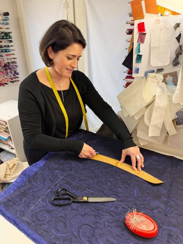 Eine Frau misst Stoff mit einem Maßband, während Schere und Stecknadeln auf dem Tisch liegen.