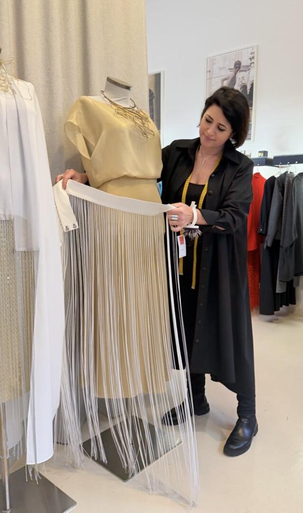 Eine Frau betrachtet ein beigefarbenes Kleid mit langen weißen Fransen an einer Schneiderpuppe in einem Modeatelier.