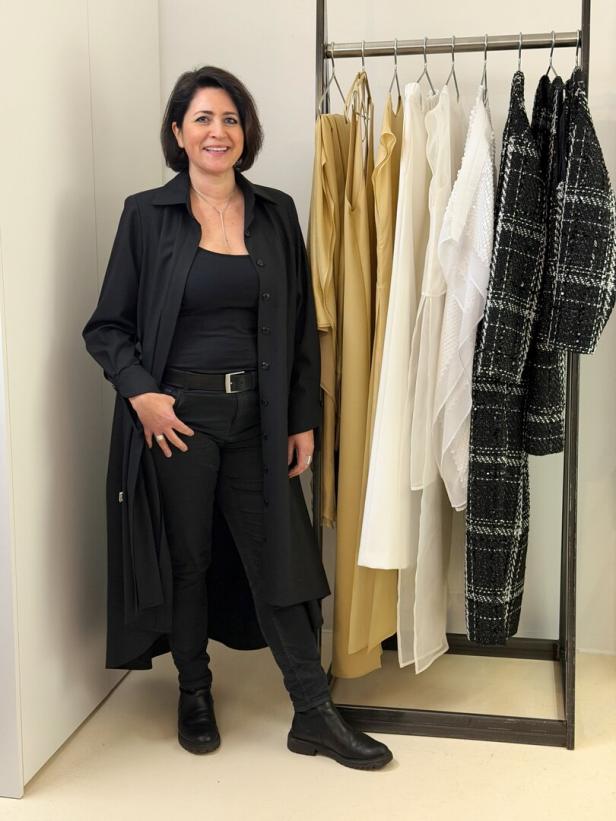 Eine Frau in schwarzer Kleidung steht neben einer Kleiderstange mit beigen, weißen und schwarz-weißen Kleidungsstücken.