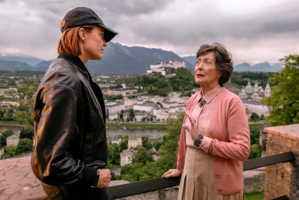 Zwei Frauen unterhalten sich im Freien mit Blick auf eine Stadt und eine Burg vor Bergkulisse.