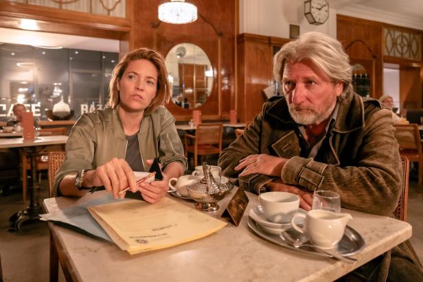 Zwei Personen sitzen an einem Tisch in einem Café, vor ihnen liegen Akten, Tassen und eine Kanne.