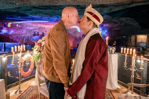 Zwei Männer küssen sich in festlicher Umgebung mit Kerzen, Blumen und romantischer Beleuchtung.