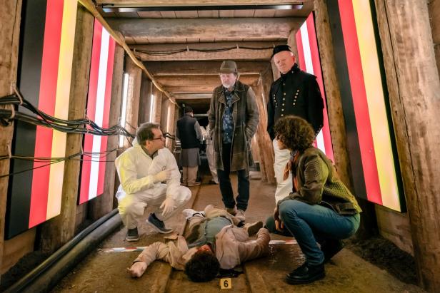 Mehrere Personen stehen und hocken in einem beleuchteten Tunnel um eine am Boden liegende leblose Person.