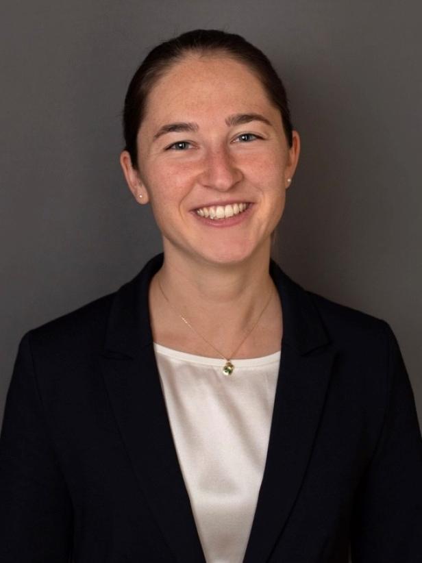 Junge Frau mit dunklen Haaren im Dutt, trägt dunklen Blazer, weiße Bluse und Kette, lächelt vor grauem Hintergrund.