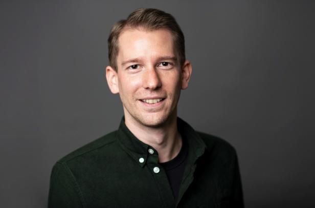 Ein Mann mit kurzen, hellbraunen Haaren trägt ein dunkles Hemd und lächelt vor grauem Hintergrund.