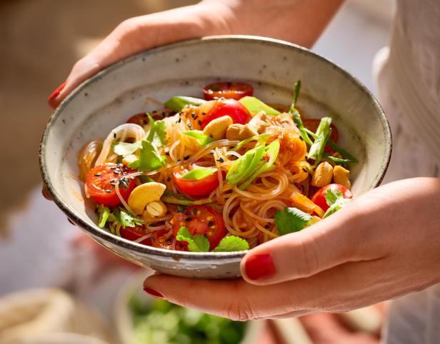 Eine Person hält eine Schale mit einem bunt angerichteten veganen Glasnudelsalat mit Kirschtomaten, Cashewkernen, Koriander und Frühlingszwiebeln.
