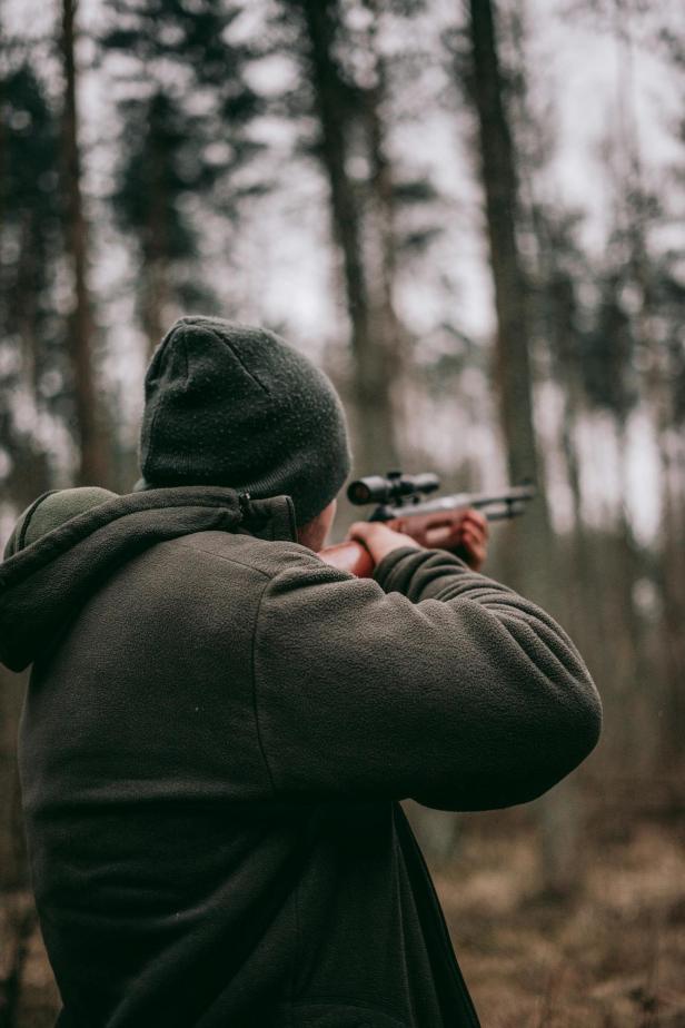 Eine Person mit Mütze und Jacke zielt mit einem Gewehr im Wald.