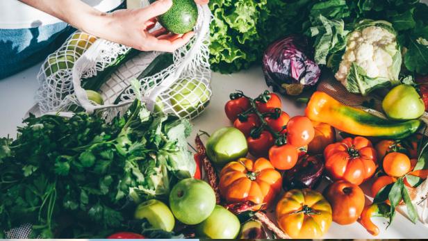 Verschiedene frische Gemüse- und Obstsorten liegen auf einem Tisch neben einem Einkaufsnetz.