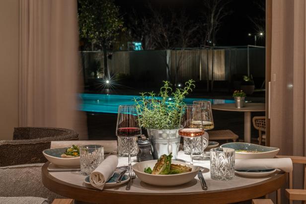 Ein elegant gedeckter Tisch mit Speisen, Weingläsern und Kräutertopf steht vor einem beleuchteten Pool bei Nacht.
