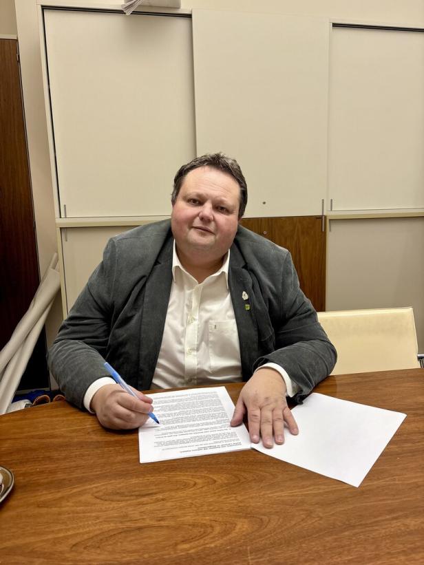 Ein Mann sitzt an einem Holztisch und unterschreibt ein Dokument mit einem blauen Stift.