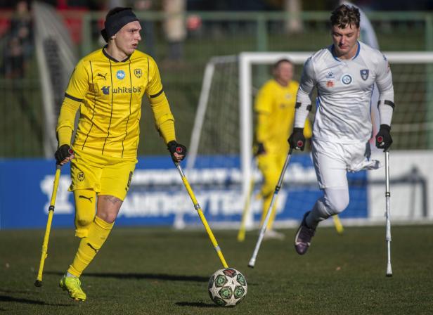 Amputierten-Fußball: Zwei Spieler mit Krücken auf dem Rasen, einer in Gelb, der andere in Weiß, spielen Fußball.