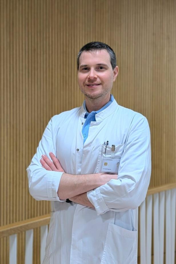 Ein Mann im weißen Arztkittel steht mit verschränkten Armen vor einer Holzvertäfelung und lächelt.