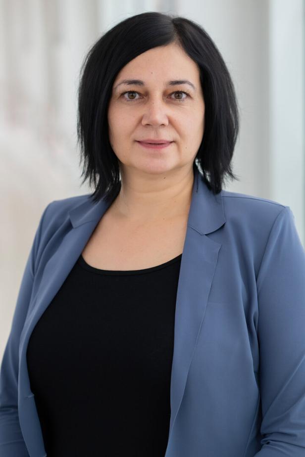 Frau mit schwarzen Haaren trägt ein schwarzes Oberteil und einen blauen Blazer, neutraler Hintergrund.