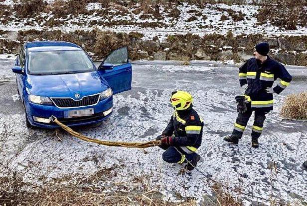 Zwei Feuerwehrleute sichern ein blaues Auto mit einem Seil auf einer vereisten Fläche.
