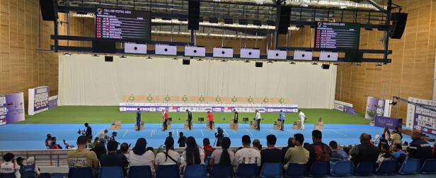 Mehrere Sportschützen stehen nebeneinander in einer Halle und zielen auf Zielscheiben, während Zuschauer im Vordergrund sitzen.