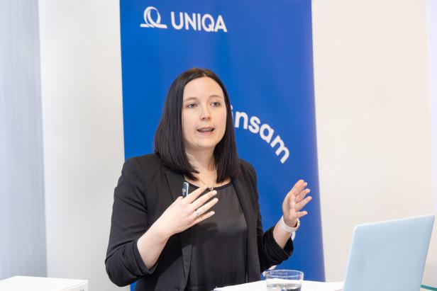 Eine Frau spricht gestikulierend vor einem blauen UNIQA-Hintergrund und einem Laptop.