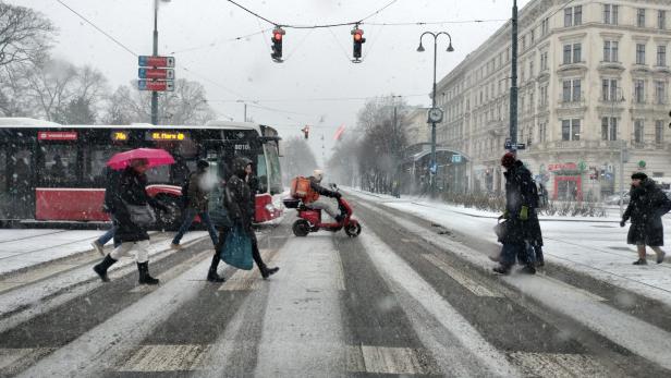 Schneefall in Wien: Menschen überqueren eine Straße, ein Bus und ein Roller sind zu sehen.