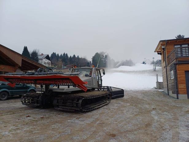 Eine Pistenraupe steht auf einem teilweise verschneiten Hang neben Gebäuden und Bäumen.