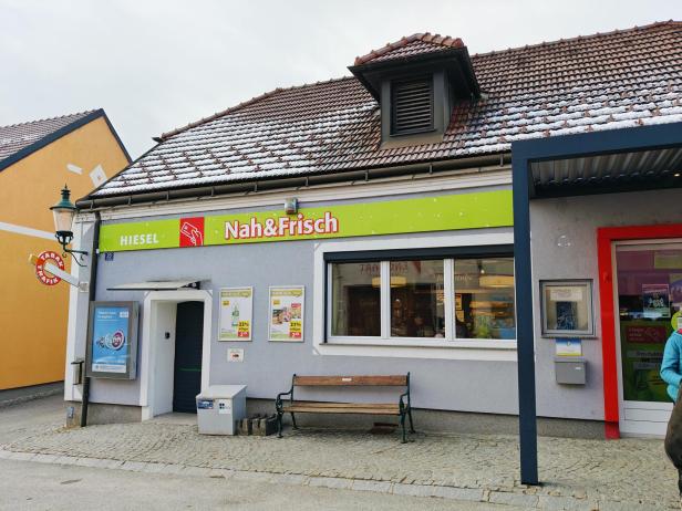 Ein kleines Geschäft mit dem Schild „Nah&Frisch“ und einer Bank davor, das Dach ist teilweise mit Schnee bedeckt.
