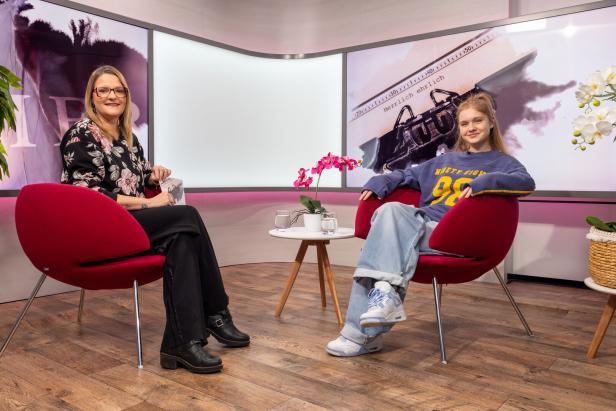 Zwei Frauen sitzen entspannt in roten Sesseln in einem modernen Studio, zwischen ihnen steht ein kleiner Tisch mit Blumen und Wasser.