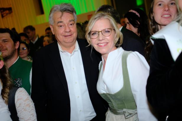 Ein Mann im Anzug und eine Frau im Dirndl lächeln gemeinsam inmitten einer festlich gekleideten Menschenmenge.