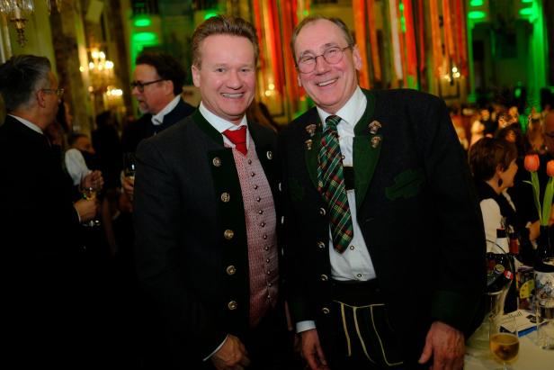 Zwei Männer in festlicher Tracht stehen lächelnd nebeneinander auf einer Veranstaltung mit festlicher Dekoration.