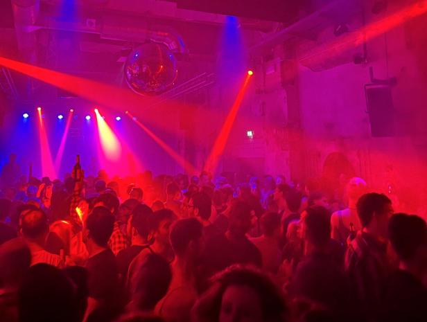 Viele Menschen tanzen in einem Club mit bunten Lichtern, Nebel und einer Discokugel an der Decke.