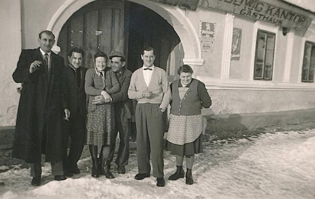 Sechs Erwachsene stehen lachend vor einem Gasthaus im Winter auf schneebedecktem Boden.