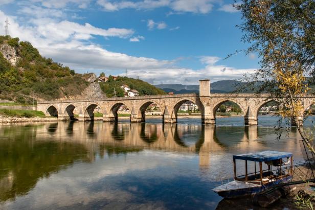 Steinerne Bogenbrücke über einen Fluss mit Bergen und Häusern im Hintergrund bei Višegrad.