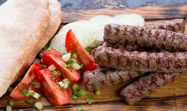 Gegrillte Ćevapčići mit Fladenbrot, Tomaten, Zwiebeln und Frühlingszwiebeln auf Holzbrett.
