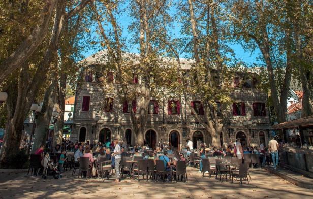 Menschen sitzen unter großen Platanen vor einem historischen Steingebäude in einem Straßencafé.