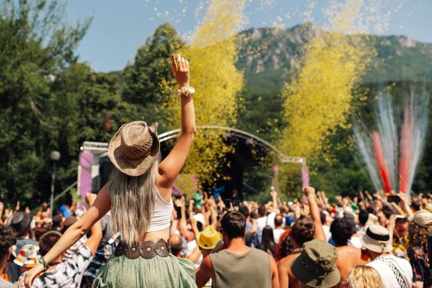 Menschen feiern bei einem Open-Air-Festival vor einer Bühne inmitten grüner Berge.