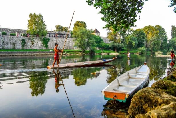 Mann stakt ein schmales Boot auf einem ruhigen Fluss, umgeben von Bäumen und Steinmauer.