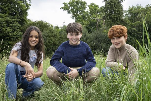 Drei Kinder hocken lachend im hohen Gras einer grünen Wiese mit Bäumen im Hintergrund.