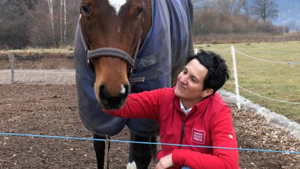 Eine Frau sitzt neben einem Pferd und streichelt dessen Hals liebevoll.