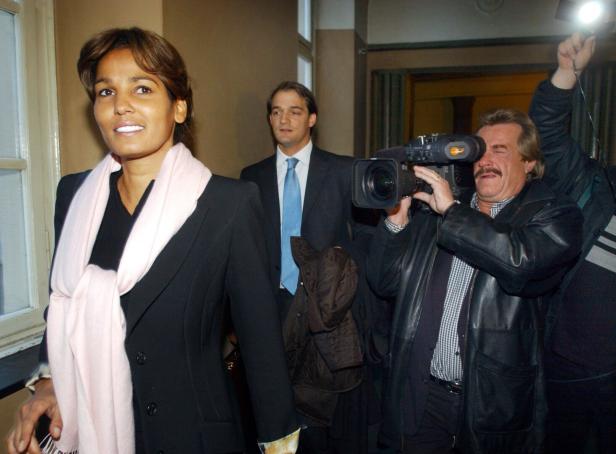 Eine Frau mit rosa Schal wird von einem Kameramann und weiteren Personen in einem Flur begleitet.