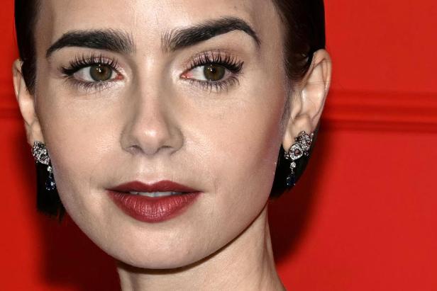 Nahaufnahme von Lily Collins mit dunklen Augenbrauen, roten Lippen und Ohrringen vor rotem Hintergrund.