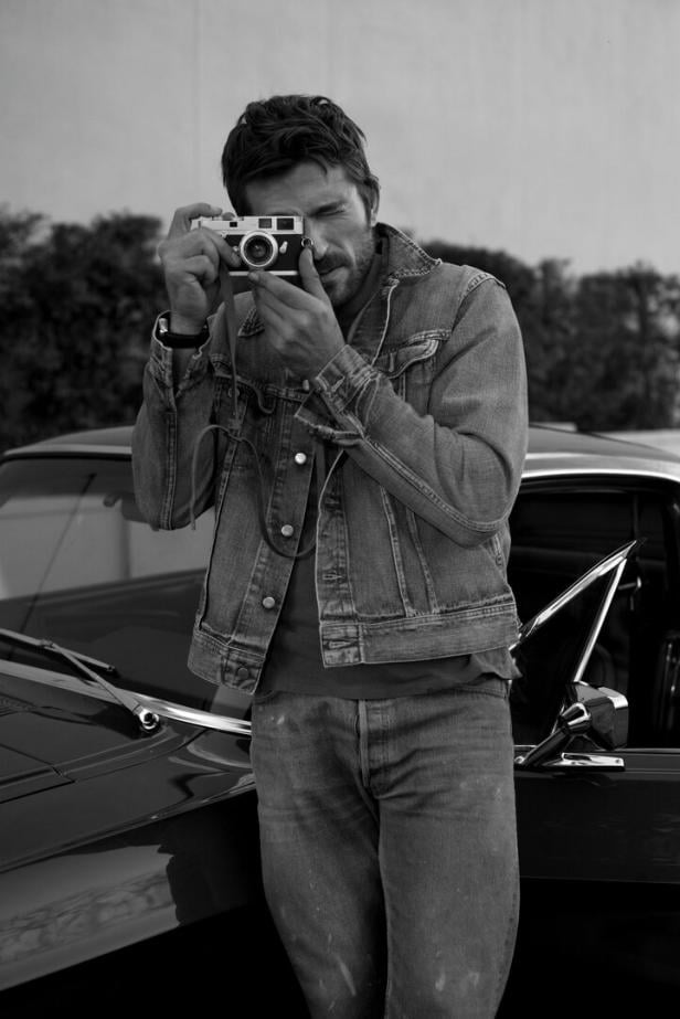 Ein Mann in Jeansjacke fotografiert mit einer Kamera und steht neben einem Auto mit geöffneter Tür.