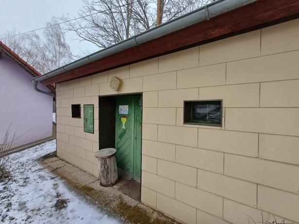 Kleines Gebäude mit grüner Tür und kleinen Fenstern, davor Schnee und ein Baumstumpf.