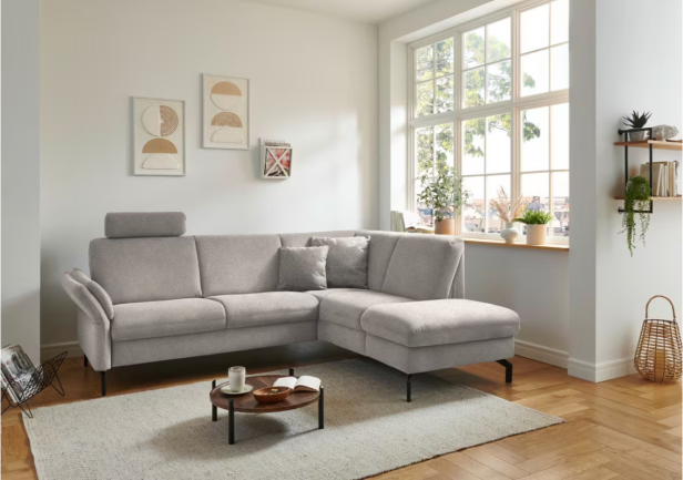 Ein hellgraues Ecksofa mit pflegeleichtem, robustem Bezug steht in einem modern eingerichteten, lichtdurchfluteten Wohnzimmer mit Pflanzen und Wandbildern.