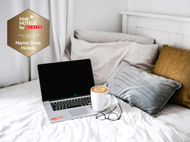 Ein Laptop, eine Tasse Kaffee und eine Brille liegen auf einem gemütlichen Hotelbett, daneben das Fine Hotels by KURIER Gütesiegel.