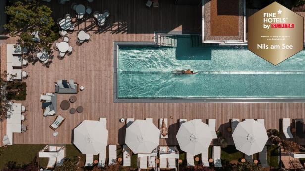 Ein Mensch schwimmt in einem rechteckigen Pool, umgeben von Liegen, Sonnenschirmen und Sitzbereichen auf einer Holzterrasse.