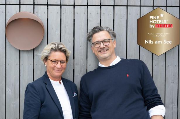 Zwei Personen mit Brillen stehen lächelnd vor einer Holzvertäfelung neben einem goldenen Hotelsiegel mit der Aufschrift „Nils am See“.