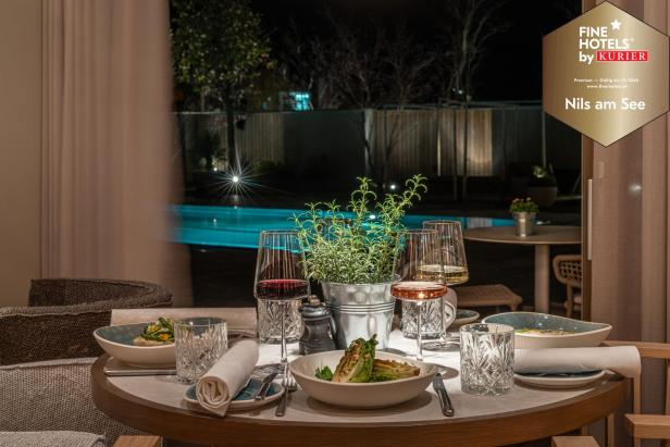 Ein elegant gedeckter Tisch mit Speisen und Weingläsern steht vor einem beleuchteten Pool im nächtlichen Garten.