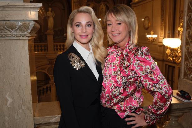 Zwei Frauen stehen nebeneinander in einem prunkvollen, goldverzierten Innenraum und lächeln in die Kamera.