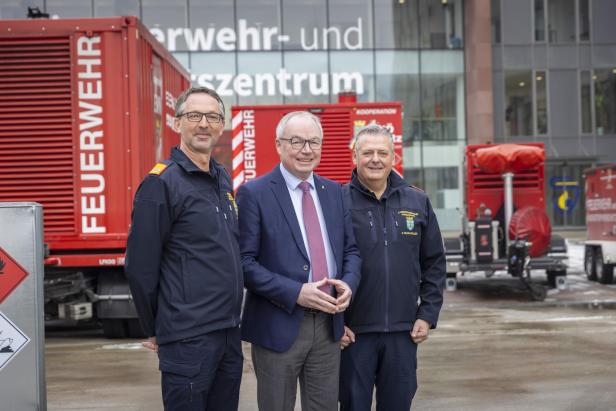 Drei Männer stehen vor roten Feuerwehrcontainern und einem modernen Gebäude mit der Aufschrift „Feuerwehr- und Katastrophenschutzzentrum“.